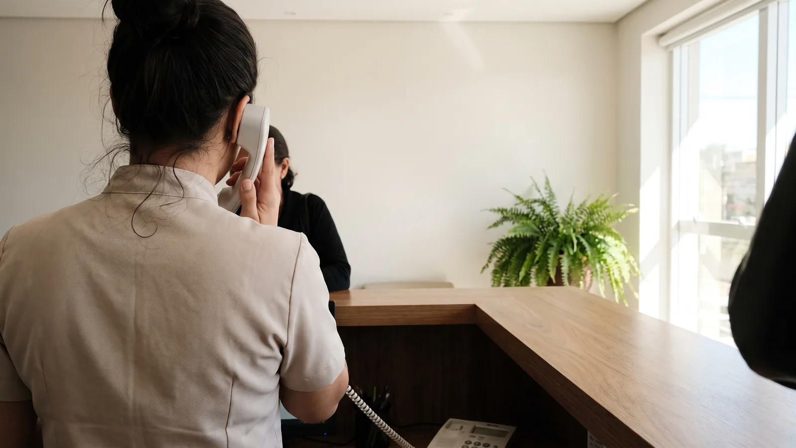 Recepcionista de clínica vista de costas atendendo o telefone no balcão, com uma paciente aguardando do outro lado e uma samambaia junto à janela